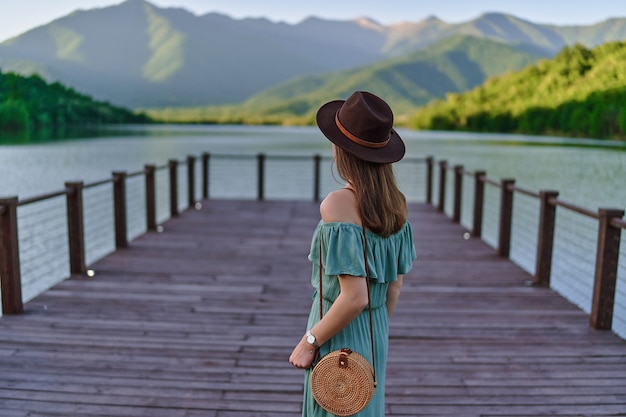 Reisendes Mädchen, das allein auf Pier steht und auf See und Berge anstarrt. Schöner Freiheitsmoment und ruhige friedliche Atmosphäre in der Natur. Rückansicht