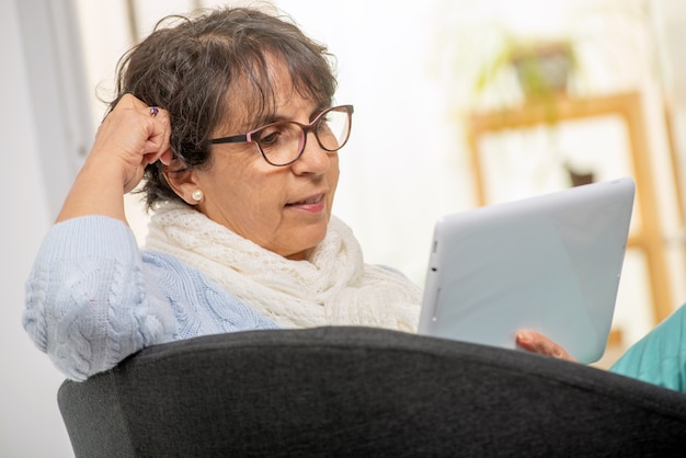 Reizend ältere Brunettefrau mit Gläsern unter Verwendung der digitalen Tablette zu Hause