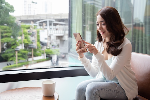 Reizend Asiatinlächeln, das gute Nachrichten am Handy während des Restes in der Kaffeestube liest