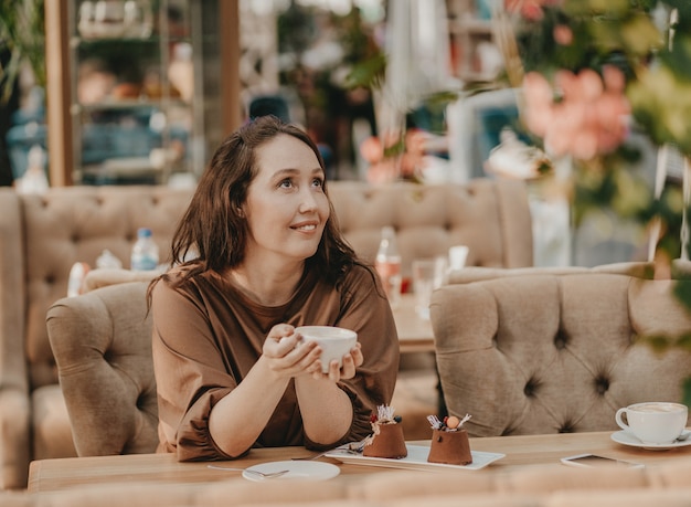 Reizend Brunettefrau mit dem langen gelockten Haar, das am Fenster im Café mit Tasse Kaffee in den Händen und im Nachtisch auf Tabelle sitzt