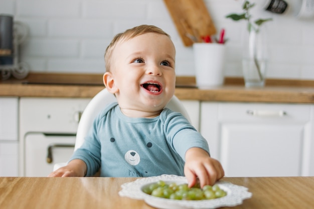 Reizend glückliches kleines baby, das zu hause erste lebensmittelgrüntraube an der hellen küche isst