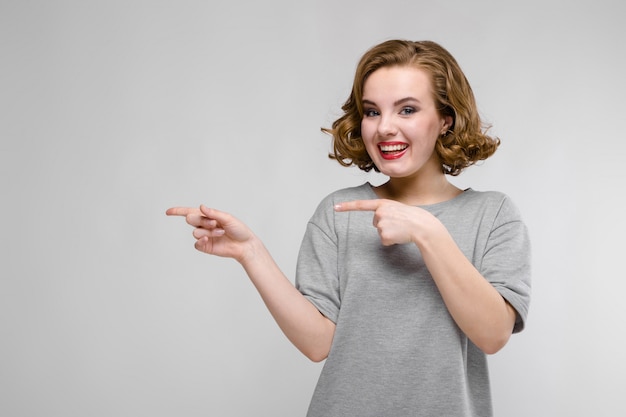 Reizend junges Mädchen in einem grauen T-Shirt auf einem grauen Hintergrund. Das Mädchen zeigt mit den Fingern zur Seite