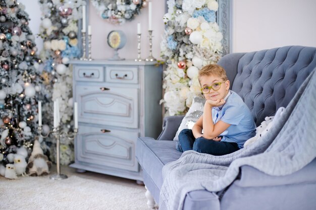 Reizend kleiner blonder Junge im blauen Hemd mit den großen Gläsern, die auf dem Sofa sitzen