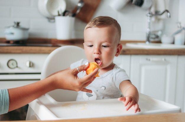 Reizend kleines Baby, das ersten Lebensmittelpfirsich an der Küche isst.