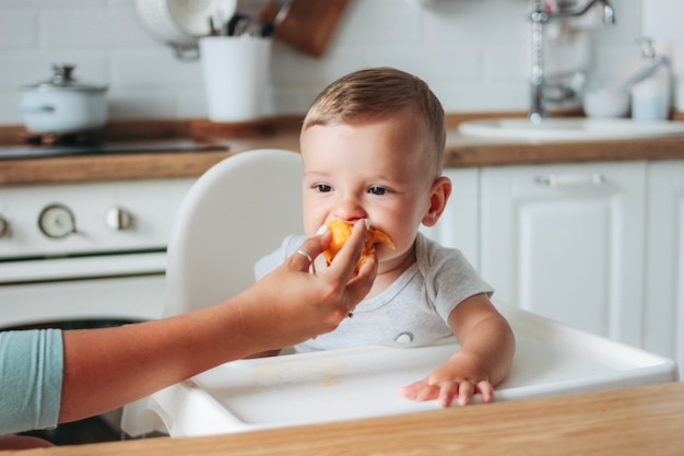 Reizend kleines Baby, das ersten Lebensmittelpfirsich an der Küche isst.