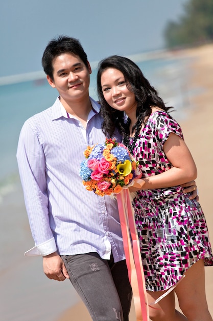 Reizende junge Paare mit dem Blumenblumenstrauß, der auf dem Strand steht