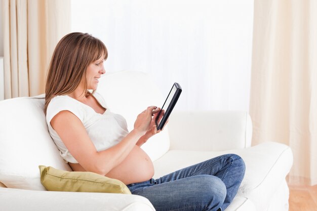 Reizende schwangere Frau, die mit einer Computertablette beim Sitzen auf einem Sofa sich entspannt