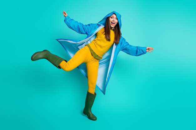 reizendes entzückendes positives Mädchen im Regenmantel, das an der blaugrünen Wand posiert