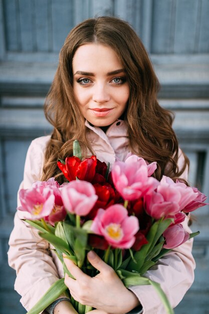 Reizendes nettes erstaunliches brunettemädchen, das vor blauer tür der alten weinlese mit einem blumenstrauß von frischen tulpen steht. frauentag. 8. märz. schöne jugendlich frau, die viele blumen als geschenk vom freund hält.