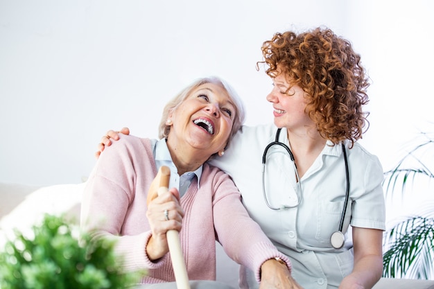 Relación amistosa entre cuidador sonriente en uniforme y feliz anciana. Joven enfermera de apoyo mirando a la mujer mayor.