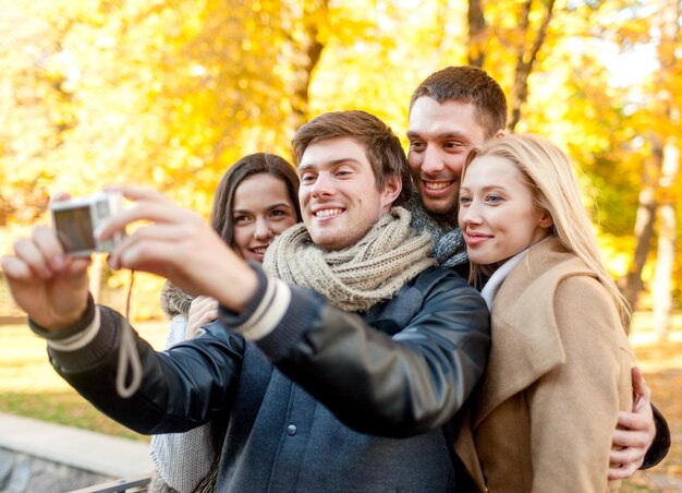 relación, temporada, amistad, tecnología y concepto de personas - grupo de hombres y mujeres sonrientes haciendo autorretrato con cámara digital en el parque de otoño