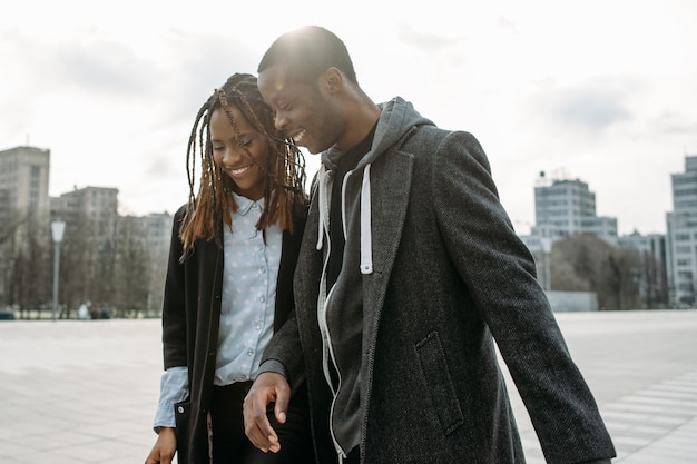 Relaciones amables. Sonriente pareja negra. Peatones afroamericanos, fondo urbano, luz solar atmosférica, estilo de moda callejera, concepto de felicidad