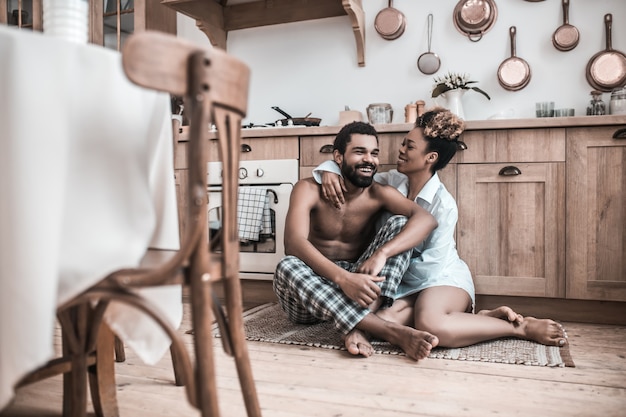 Relaciones familiares. Alegres afroamericanos esposo y esposa sentados en una alfombra en el piso abrazándose comunicarse