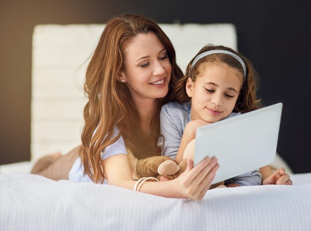 Relaja la tableta y la madre con la niña en la cama en casa mira video en línea en la aplicación internet o redes sociales Sonríe amor y madre con el niño transmisión de películas o espectáculos en tecnología digital en el dormitorio