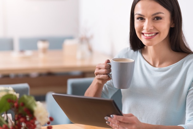 Relajación deliciosa. Hermosa mujer encantadora alegre sonriendo y tomando una bebida caliente mientras usa su gadget en una cafetería.