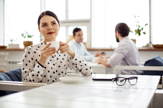 Relajación. Mujer joven de pelo oscuro bastante meditativa sonriendo y tomando una taza de café mientras está sentado en la mesa y pensando