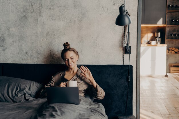 Relajada mujer joven tranquila acostada en la cama en casa con una taza de café por la mañana mientras charla en línea con sus amigos en la computadora portátil, sonriendo y saludándoles con la mano, vestida con un acogedor pijama de satén