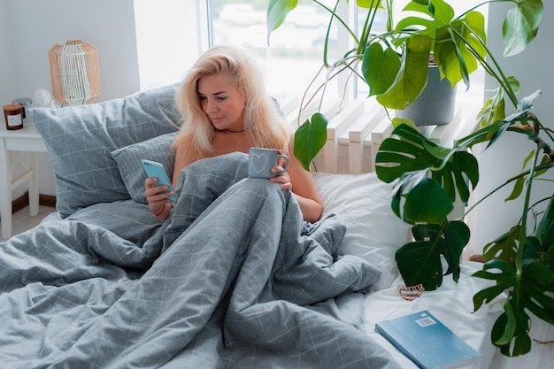 Relajada mujer rubia positiva de mediana edad tomando selfie en el teléfono móvil mientras se relaja en la cama en casa