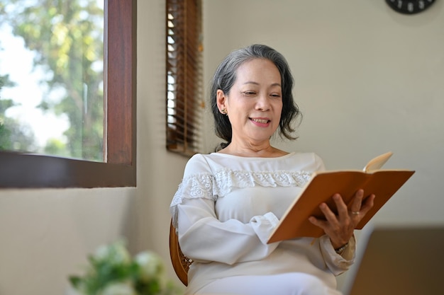 Relajada y tranquila mujer asiática jubilada de los años 60 leyendo un libro en el salón de su casa
