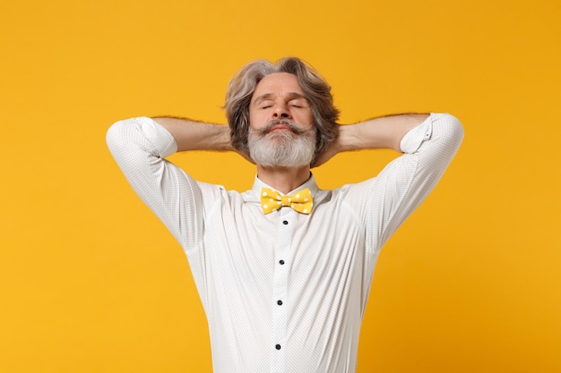 Relajado anciano de pelo gris bigote barbudo hombre con camisa blanca, pajarita posando aislado sobre fondo de pared naranja amarillo. Concepto de estilo de vida de las personas. Simulacros de espacio de copia. Dormir con las manos detrás de la cabeza.