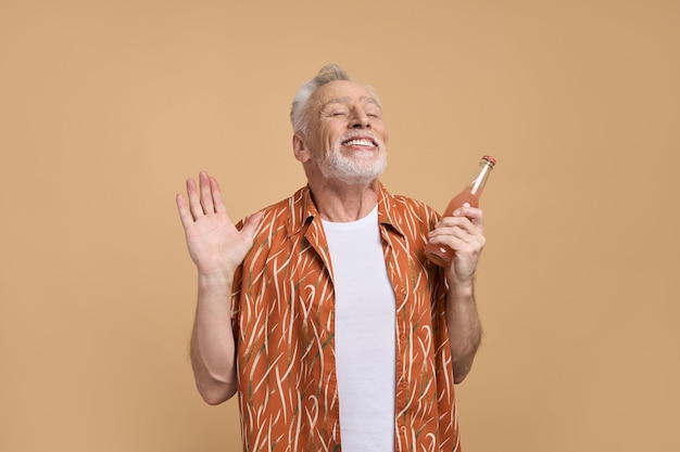 Relajado anciano sonriente sosteniendo refrescante limonada posando con los ojos cerrados sobre fondo beige