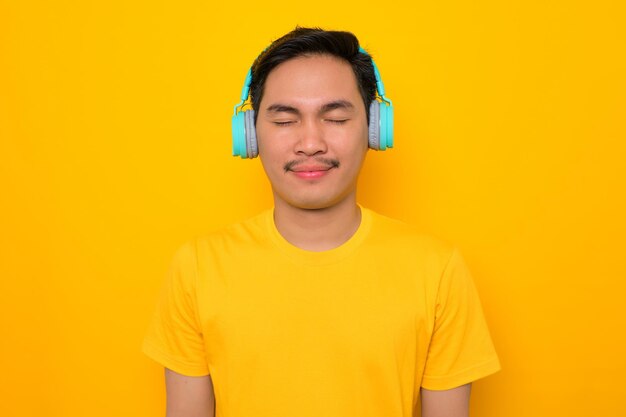 Relajado joven asiático en camiseta casual escuchando música en auriculares aislados sobre fondo amarillo Personas emociones concepto de estilo de vida