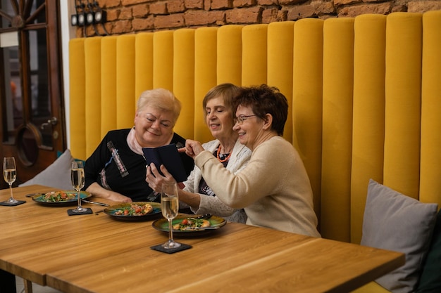 Relajando a mujeres mayores de cabello oscuro y rubio mirando el teléfono, comiendo pizza en platos, discutiendo fotos mientras celebran su reunión o vacaciones en un café moderno con paredes naranjas