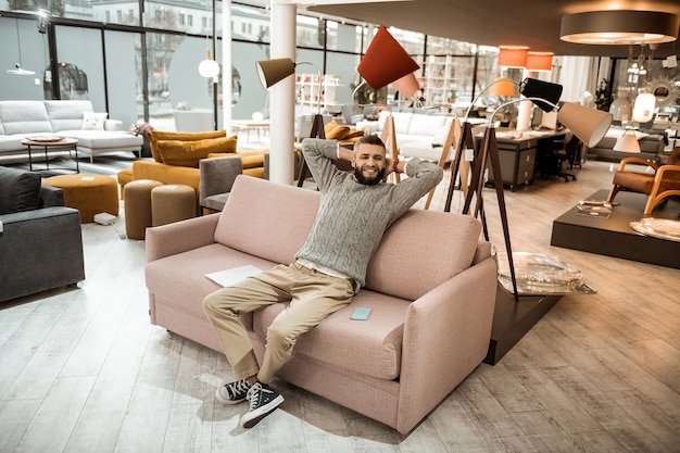 Relajándose en el sofá. Hombre sonriente en suéter gris descansando en el sofá en medio de la tienda de muebles