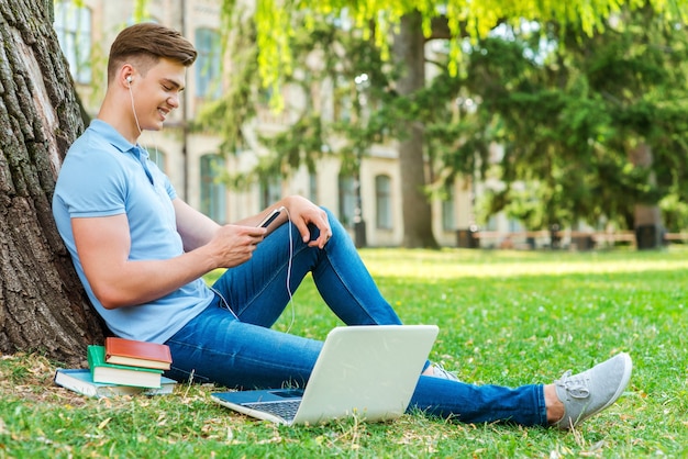 Relajarse después de clase. Vista lateral del guapo estudiante universitario masculino escuchando un reproductor de MP3 mientras está sentado en el césped y apoyado en el árbol