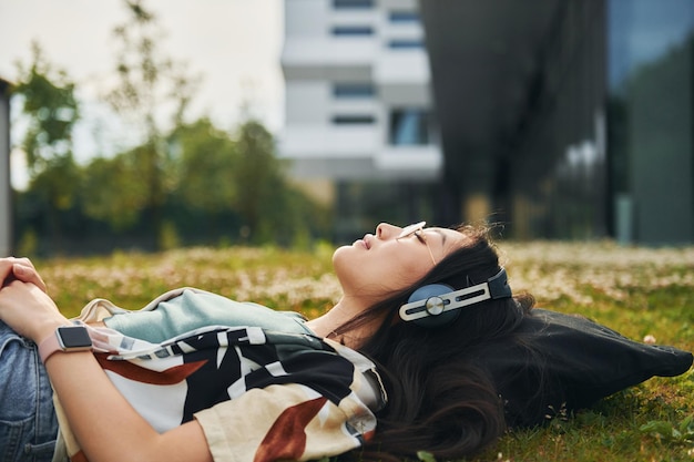 Relajarse en la hierba En los auriculares La mujer asiática joven está al aire libre durante el día