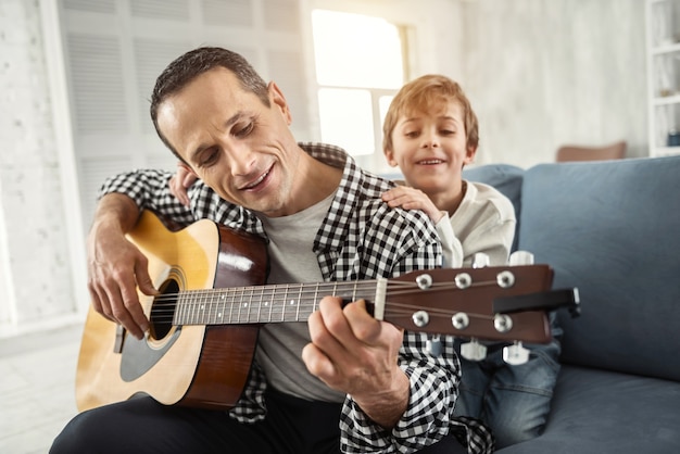 Relajarse juntos. Hombre de pelo oscuro alerta atractivo sonriendo y tocando la guitarra y su hijo abrazándolo