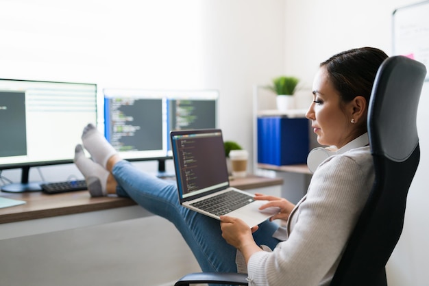 Relajarse durante mi trabajo de oficina en casa. Atractiva mujer latina poniendo sus pies encima de su escritorio y escribiendo código en su computadora portátil