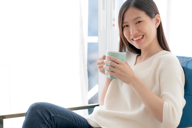 Relájese y apacible mujer asiática disfrute de un fin de semana de vacaciones con té caliente y un buen libro por la mañana cerca del fondo de la casa de la ventana