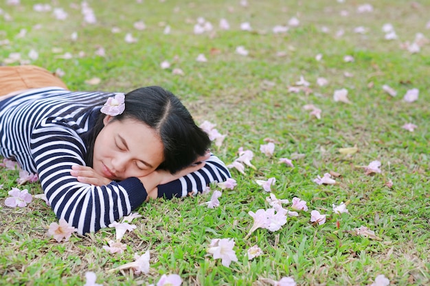 Relájese y duerma a la mujer asiática joven que miente en campo verde con completamente la flor rosada de la caída en el jardín al aire libre.