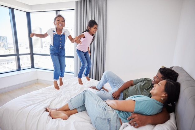 Relájese, la familia feliz y los niños saltan sobre el colchón de la cama en la casa de Filipinas con los padres mirando Los niños emocionados y felices disfrutan de la diversión junto con mamá y papá en un dormitorio acogedor