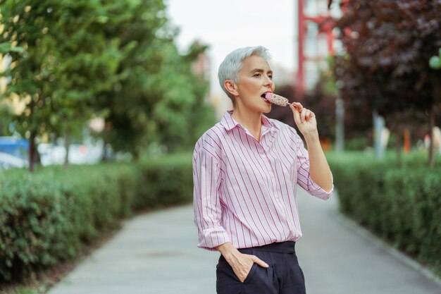 Relaxada feliz mulher de cabelos grisalhos comendo sorvete saboroso e olhando para longe de pé no parque férias de verão conceito de estilo de vida positivo