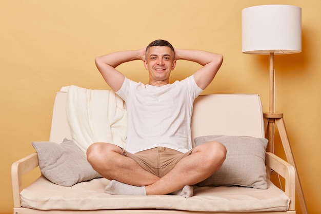 Relaxado encantado moreno homem adulto vestindo roupas casuais sentado no sofá contra a parede bege descansando em casa mantém as mãos atrás da cabeça