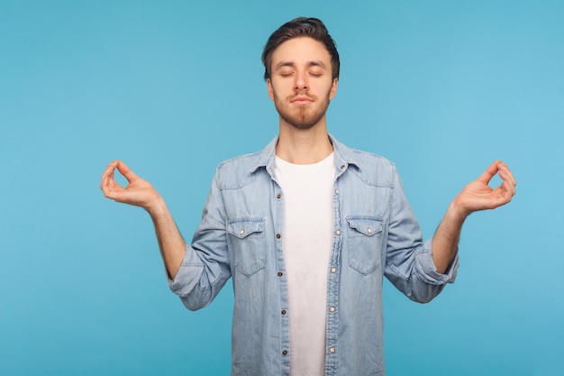 Relaxamento da mente pacífica Retrato de homem calmo na camisa jeans do trabalhador em pé com os olhos fechados e gesto de mudra meditando concentrando pensamentos em estúdio interior isolado em fundo azul