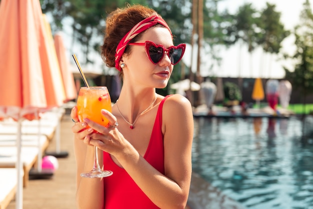 Relaxamento maravilhoso. mulher jovem e bonita olhando para a piscina enquanto segura um coquetel nas mãos