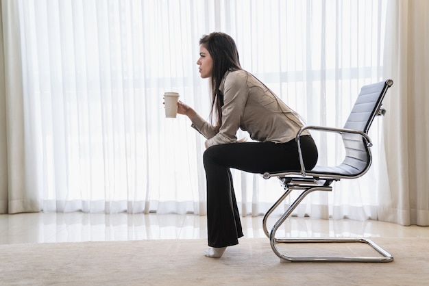 Relaxamento matinal. Mulher latina sentada na poltrona e tomando café contra a janela. Espaço livre.