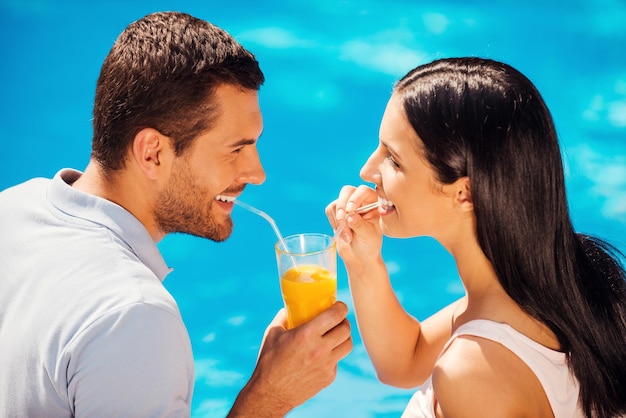 Relaxando à beira da piscina juntos. Vista superior de um lindo casal jovem em trajes casuais bebendo coquetel em um copo e sorrindo enquanto sentam-se à beira da piscina