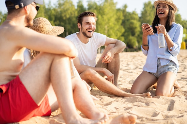 Relaxando com melhores amigos. Grupo de jovens felizes sentados juntos na praia conversando e bebendo cerveja