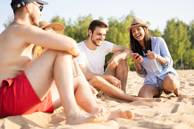 Relaxando com melhores amigos. Grupo de jovens felizes sentados juntos na praia conversando e bebendo cerveja