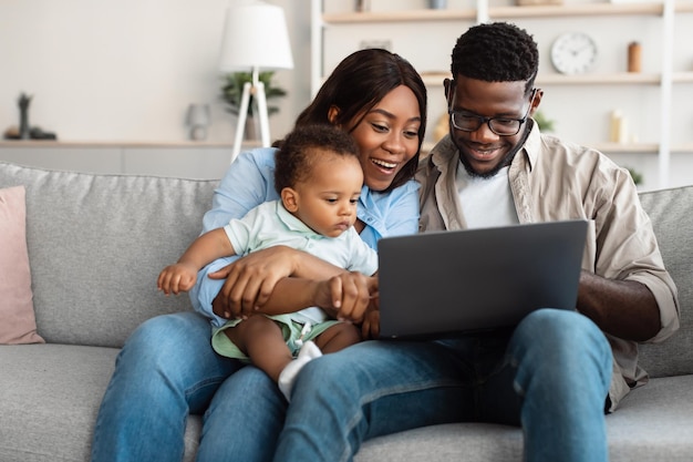 Relaxando Juntos. Retrato de pais afro-americanos felizes e seu filho fofo usando laptop, passando um tempo juntos, assistindo filme ou navegando na internet, sentados no sofá da sala de estar