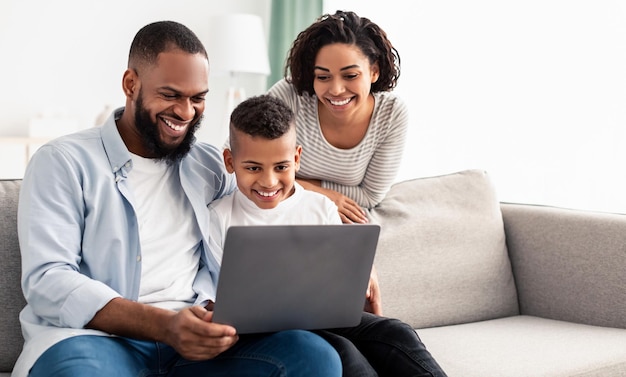 Foto relaxando juntos. retrato de pais negros felizes e seu filho pré-adolescente usando laptop, passando um tempo juntos, assistindo filme ou navegando na internet, sentados no sofá da sala de estar, copie o espaço, banner