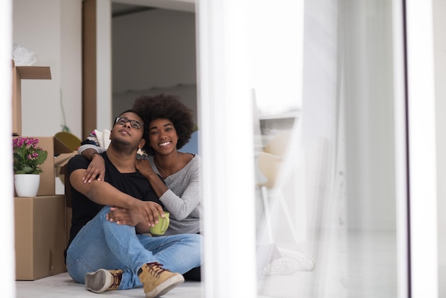 Foto relaxando na casa nova. alegre jovem casal afro-americano sentado no chão e bebendo café enquanto caixas de papelão ao seu redor