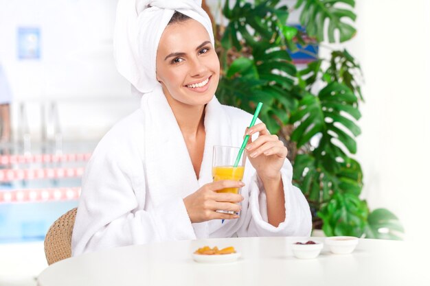 Relaxando no spa. Mulher jovem e atraente em roupão de banho segurando um copo com suco e sorrindo enquanto está sentada em frente à piscina