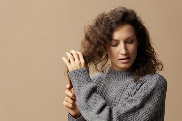 Foto relaxante mulher bonita muito encaracolada na camisola casual cinza com cabelo pentes de escova de cabelo parece de lado posando isolado sobre fundo pastel bege conceito de cabelo danificado indisciplinado problemático copie o espaço