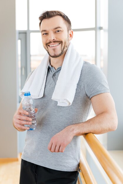 Relaxar após o treinamento esportivo. Jovem alegre com uma toalha nos ombros, segurando uma garrafa com água e sorrindo em pé no clube esportivo