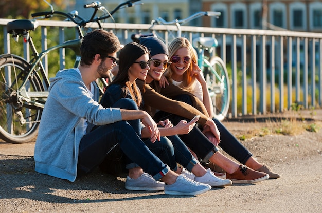 Relaxar após um dia de passeios. Grupo de jovens sorrindo, unindo-se uns aos outros e olhando para o telefone inteligente enquanto estão sentados ao ar livre com bicicletas ao fundo